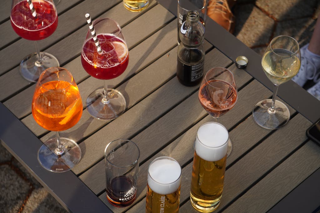 A variety of colorful drinks including cocktails, wine, and beer are arranged on a wooden table outdoors.