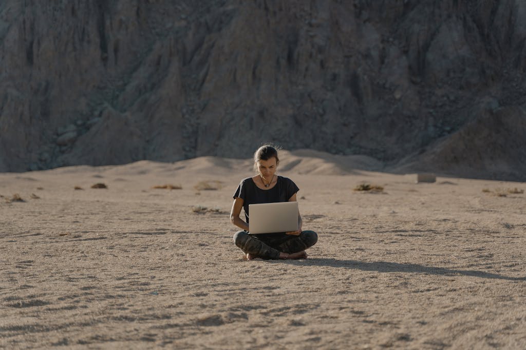 A woman works remotely on a laptop while sitting alone in a sandy desert under the sunlight.
