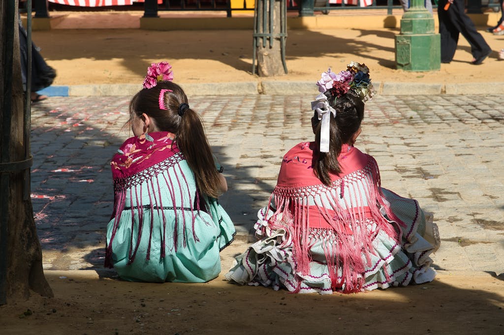 Girls in traditional flamenco dresses sitting on sidewalks during Seville's festive Feria.
