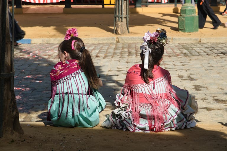 Girls in traditional flamenco dresses sitting on sidewalks during Seville's festive Feria.