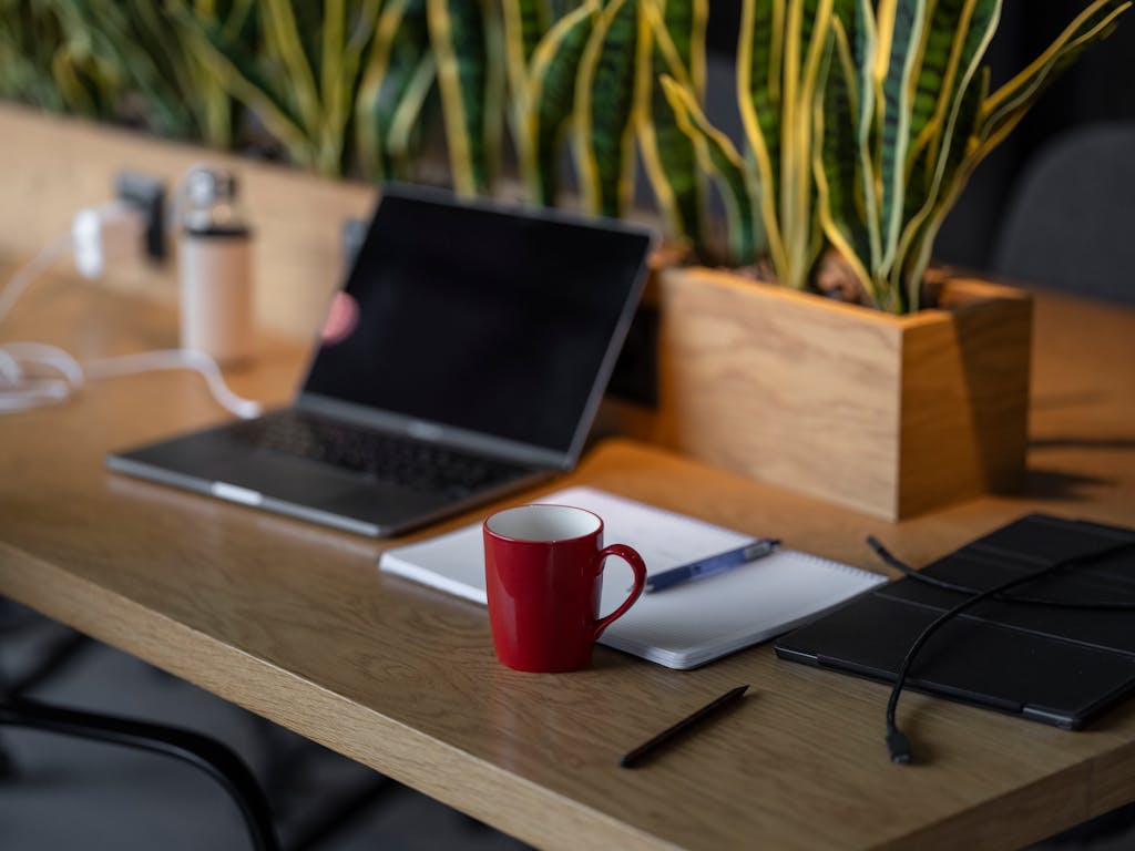 A cozy indoor workspace featuring a laptop, red coffee mug, notebooks, and plants.